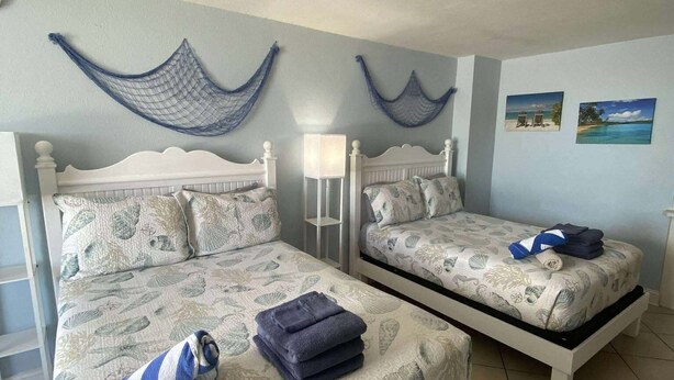 A bedroom with two beds, each with a white frame and a nautical-themed bedspread. There are two wall-mounted hammocks and two framed pictures on the wall. - Charming Studio condo in excellent Daytona Beach (Daytona Beach)