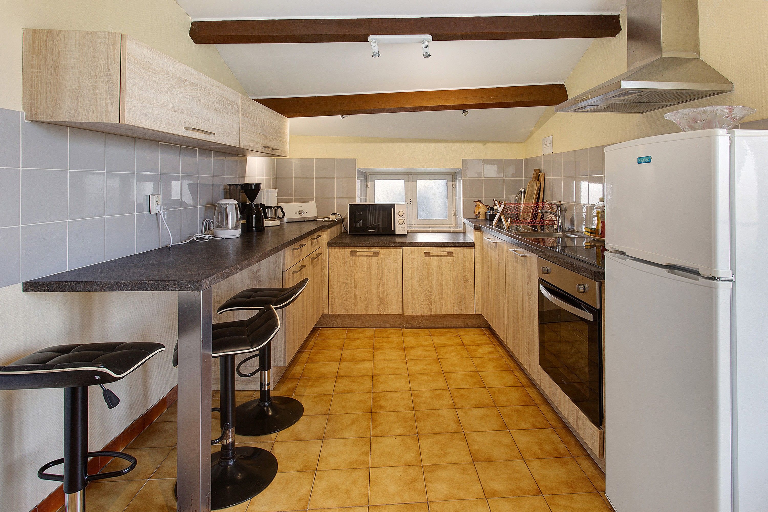 A compact kitchen with a microwave, coffee maker, and a refrigerator. There are two stools and a window above the sink.