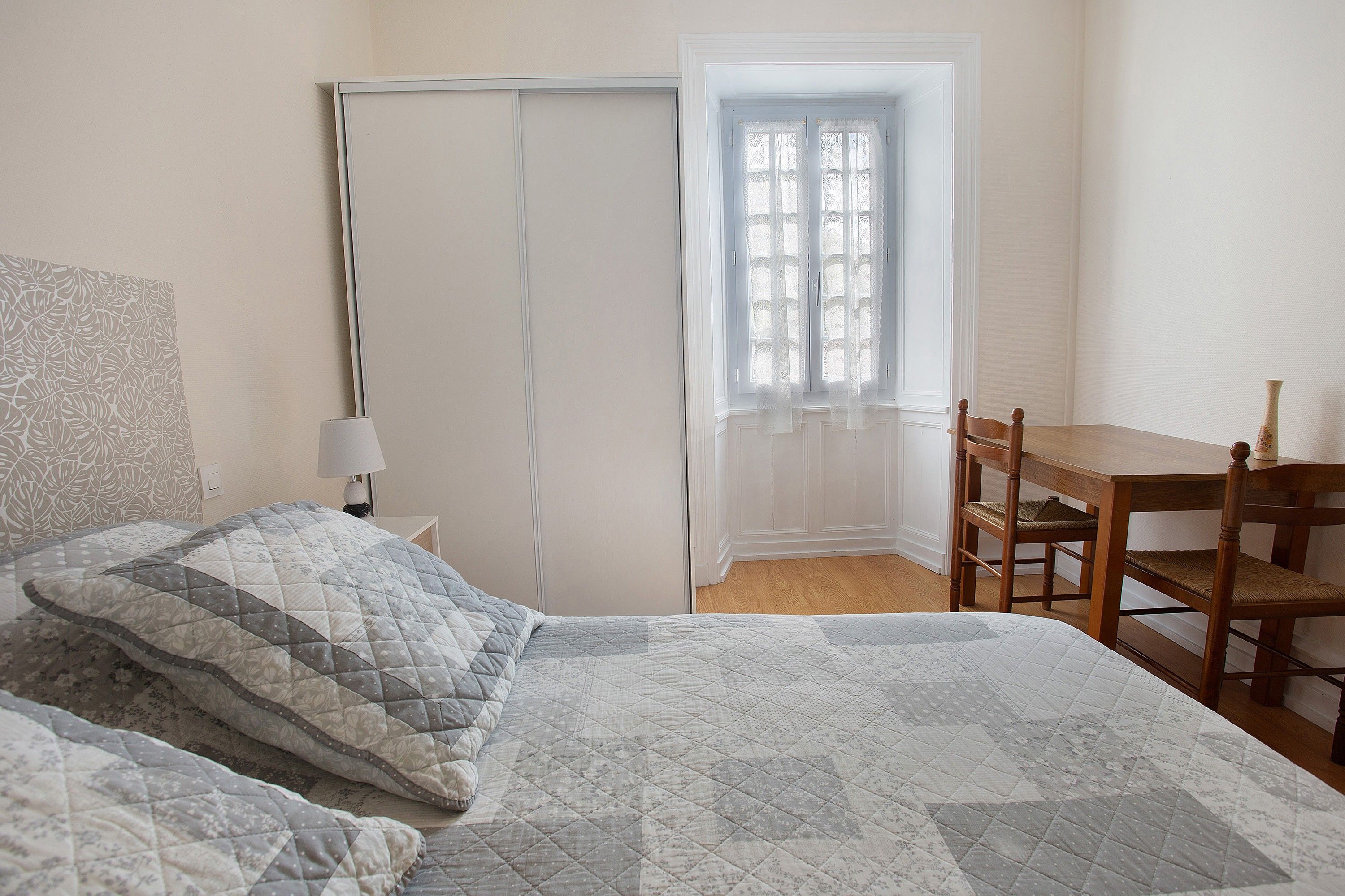 Ein Schlafzimmer mit einem Bett, einem Sessel, einem Tisch, einem Fenster mit Spitzenvorhängen und einer Tür.