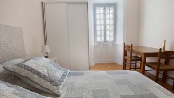 Ein Schlafzimmer mit einem Bett, einem Sessel, einem Tisch, einem Fenster mit Spitzenvorhängen und einer Tür.