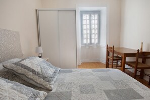 Ein Schlafzimmer mit einem Bett, einem Sessel, einem Tisch, einem Fenster mit Spitzenvorhängen und einer Tür.