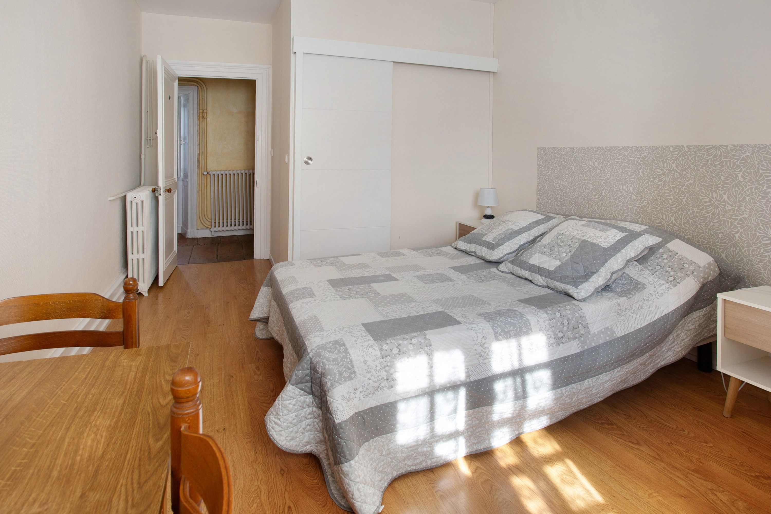 Une chambre à coucher comprenant un lit, une chaise, une table de chevet, un radiateur et une porte donnant sur une autre pièce.