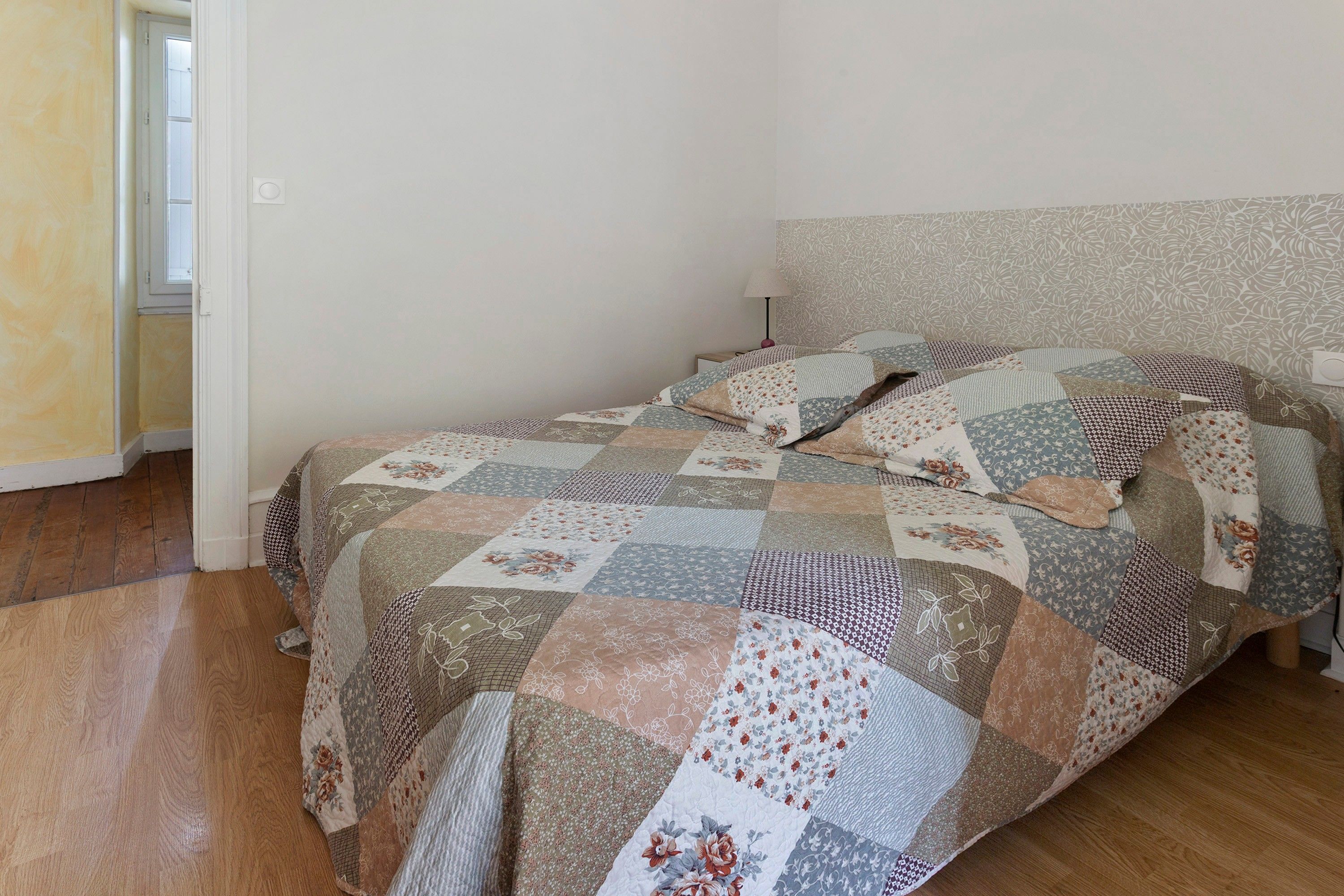 Une chambre avec un plancher en bois, un lit recouvert d’une courtepointe à motifs et un mur uni.