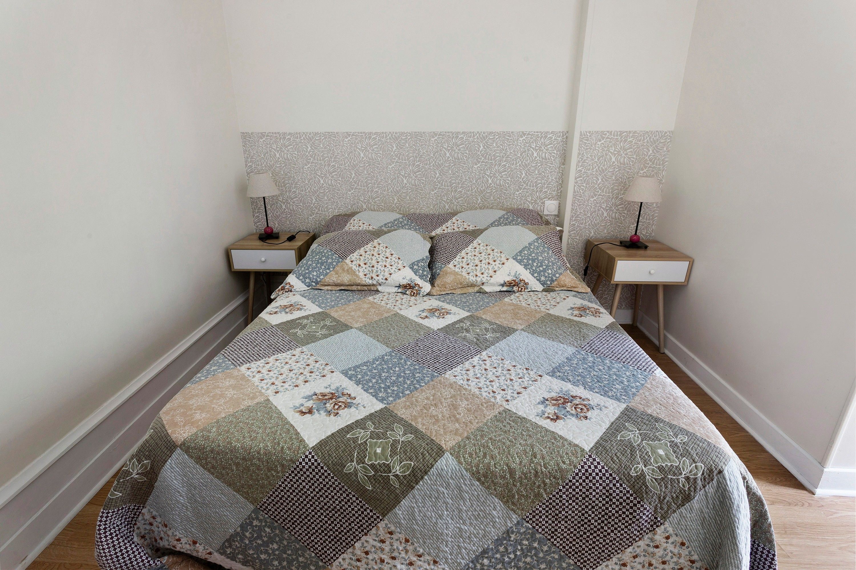 Una cama con colcha de retazos, dos mesitas de noche con lámparas y papel tapiz estampado.