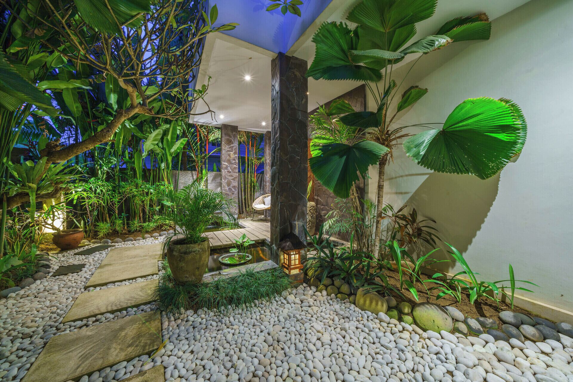 Un jardín interior bien iluminado con diversas plantas, un sendero de piedra y una zona de estar con un farol.