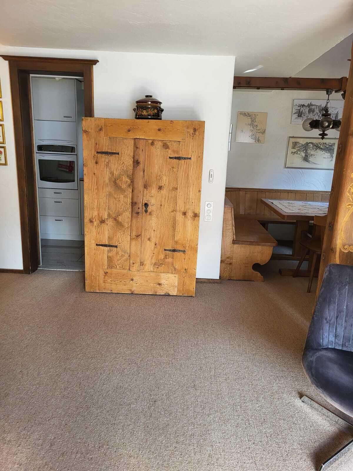 A room with a wooden wardrobe, a dining table with chairs, and a kitchenette with a built-in oven.