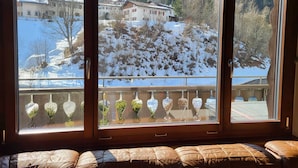 Et værelse med et stort vindue der udover et snelandskab, en balkon med dekorative plantekasser, og en polstret bænk.