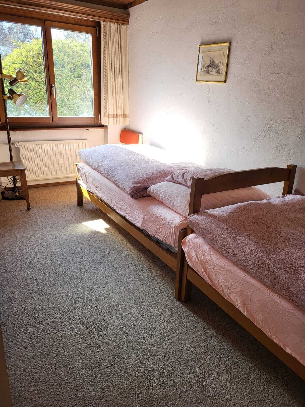 A bedroom with two single beds, a desk, a chair, a radiator, and a window with curtains.