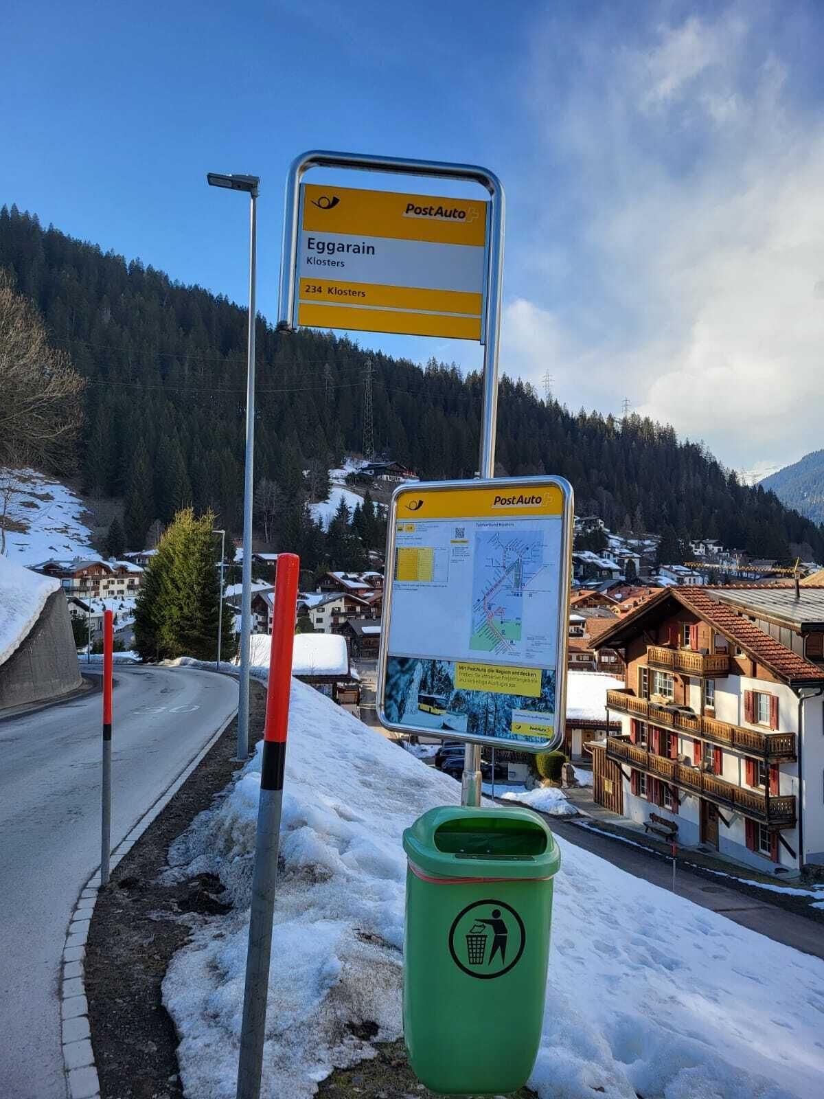A signpost with directions to 'Eggarain Klosters' and a map, a green trash bin, and a snow-covered landscape with buildings.