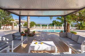 A covered outdoor seating area with a pool, white furniture, and a view of the sea.