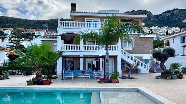 Un espace aménagé au bord de la piscine avec un fauteuil de relaxation, un bâtiment à deux étages doté de balcons et un paysage montagneux en arrière-plan.