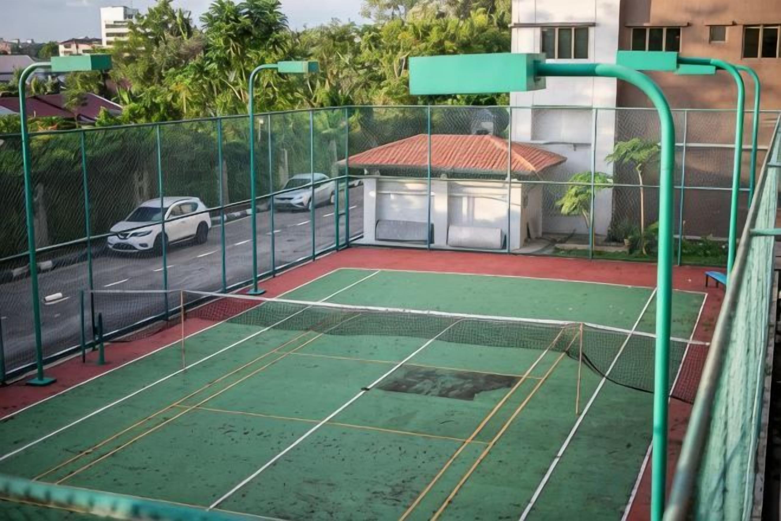 Una cancha de tenis exterior con superficie verde, rodeada por una valla verde y contigua a una calle con coches estacionados.