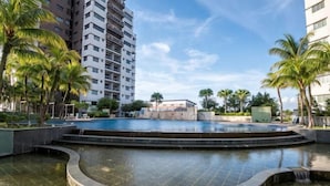 Une piscine agrémentée d’une cascade, entourée de palmiers et de bâtiments modernes.