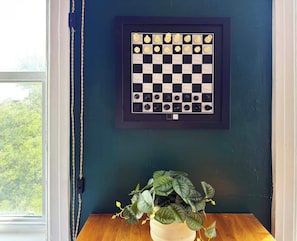 A chessboard framed on a dark wall, a potted plant on a wooden table, and a window with curtains.