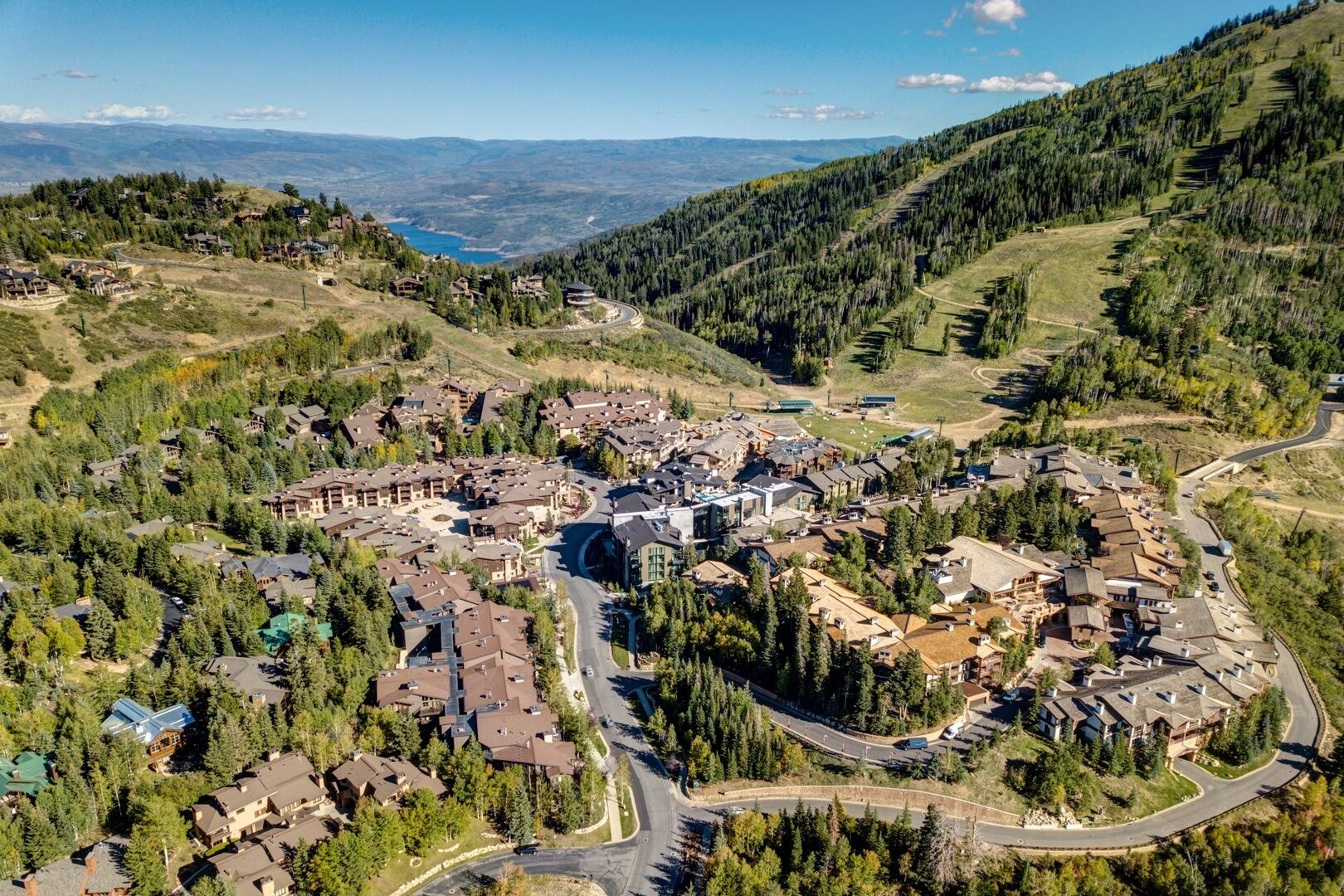 Aerial view of mountain resort village nestled in scenic valley with forested hillsides and distant lake views.