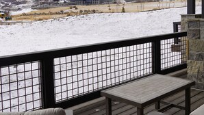 A wooden deck with outdoor seating, overlooking a snowy mountain landscape with ski slopes and buildings.