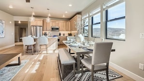 A modern kitchen with wooden cabinets, a dining area with a large table and chairs, and a view of the landscape through large windows.