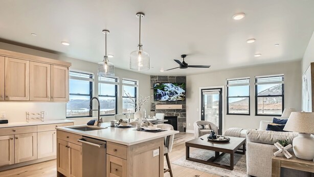 A modern kitchen with wooden cabinets, a central island, and pendant lighting. A dining area with a sofa and coffee table is visible.