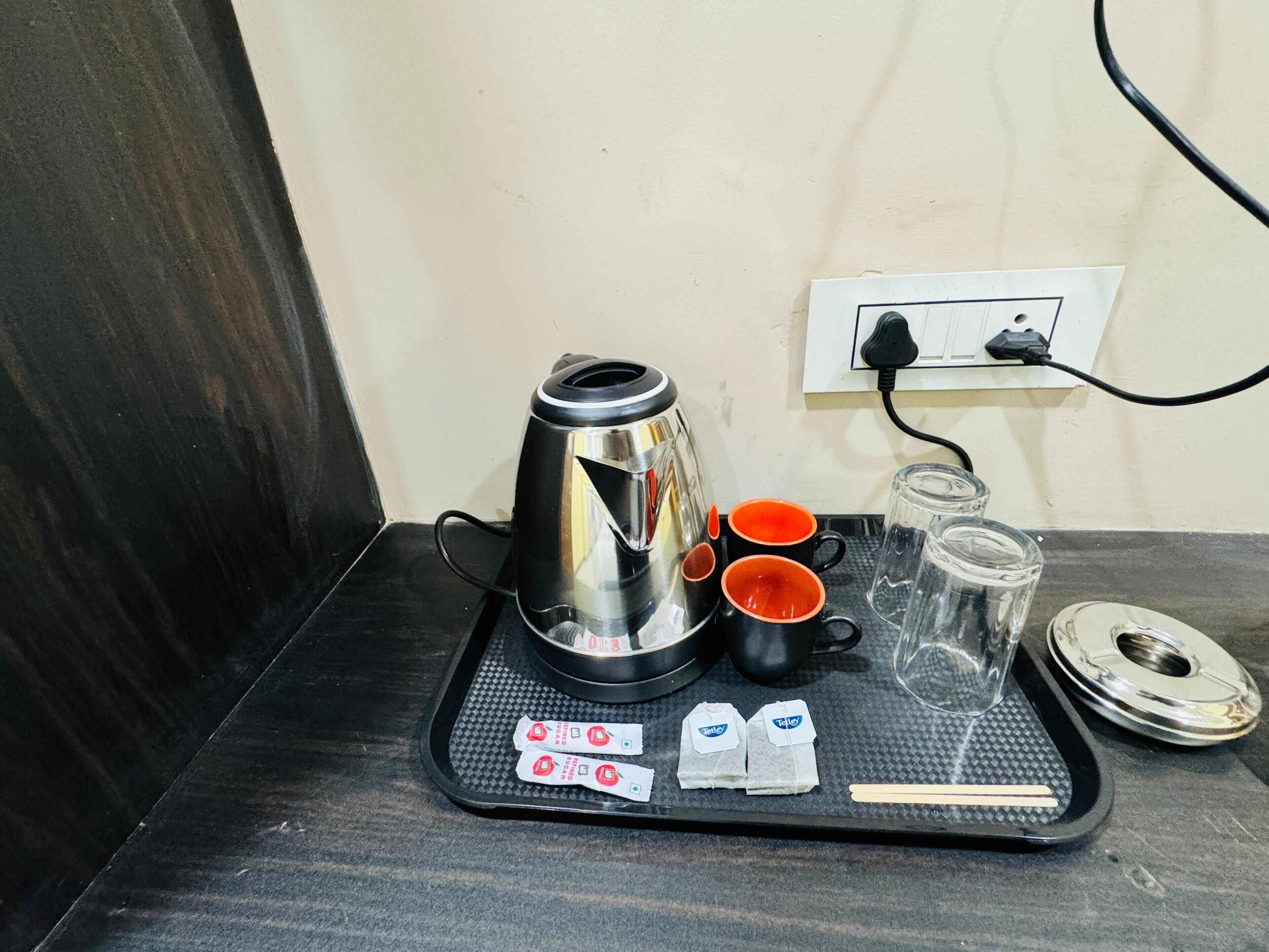 Una estación de café con un hervidor, dos tazas anaranjadas, un vaso y bolsitas de té sobre una bandeja.