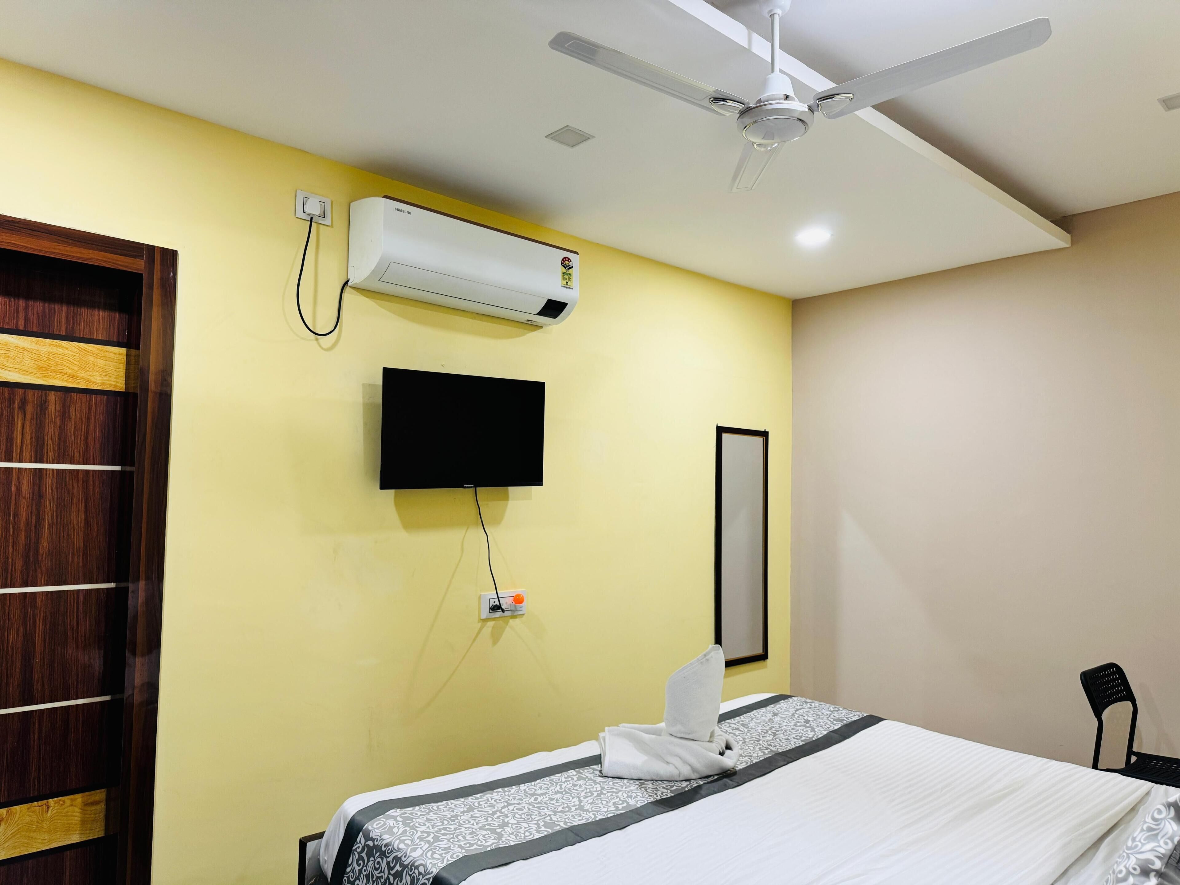 A hotel room with a bed, a flat-screen TV, an air conditioning unit, a ceiling fan, and a wooden door.