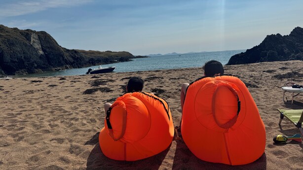 To personer sidder på orange strandstole og ser ud over havet, med en båd i vandet og klipper i baggrunden.