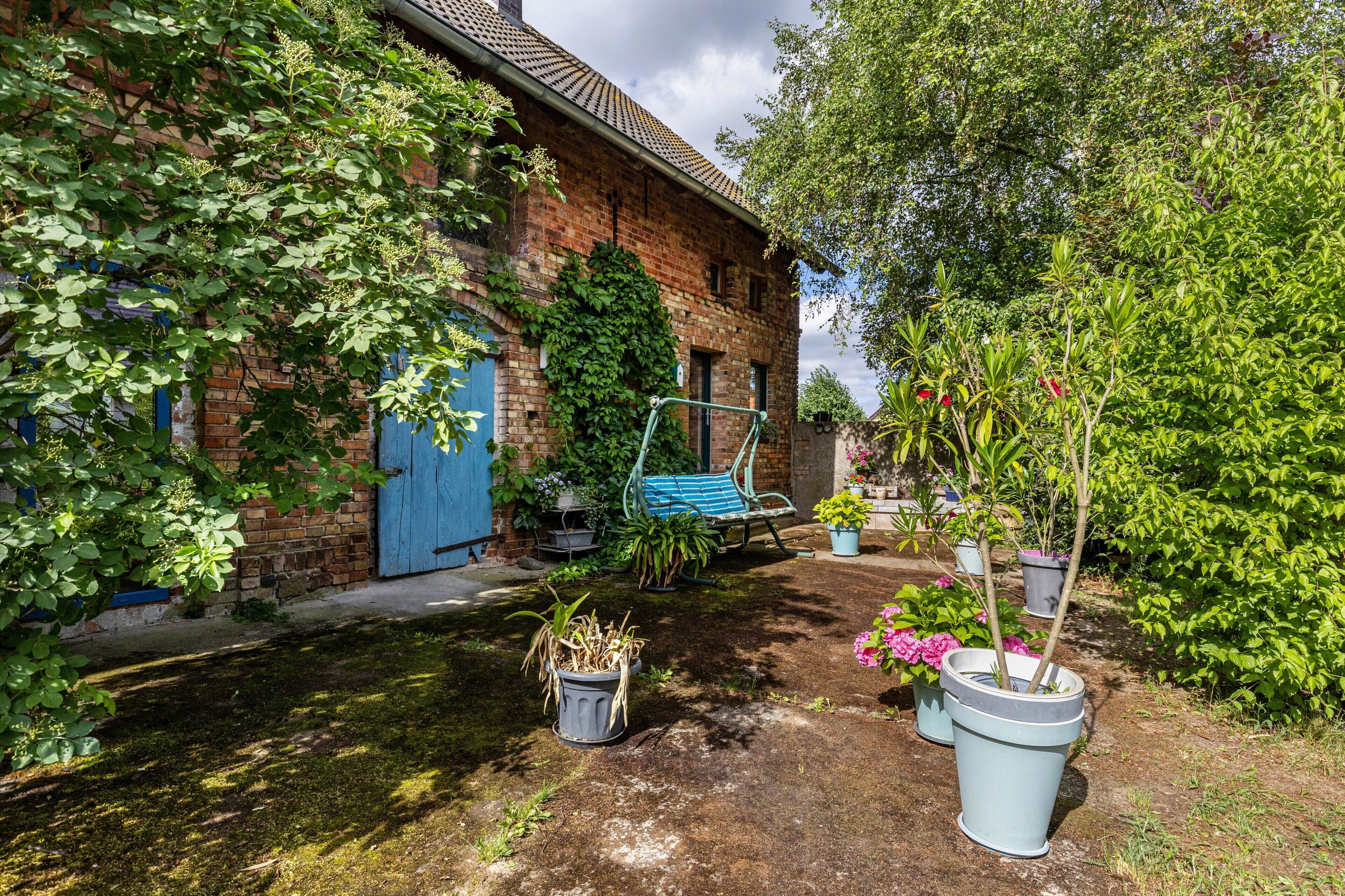 Un bâtiment en briques avec une porte bleue, un jardin agrémenté de plantes en pot et une chaise bleue.