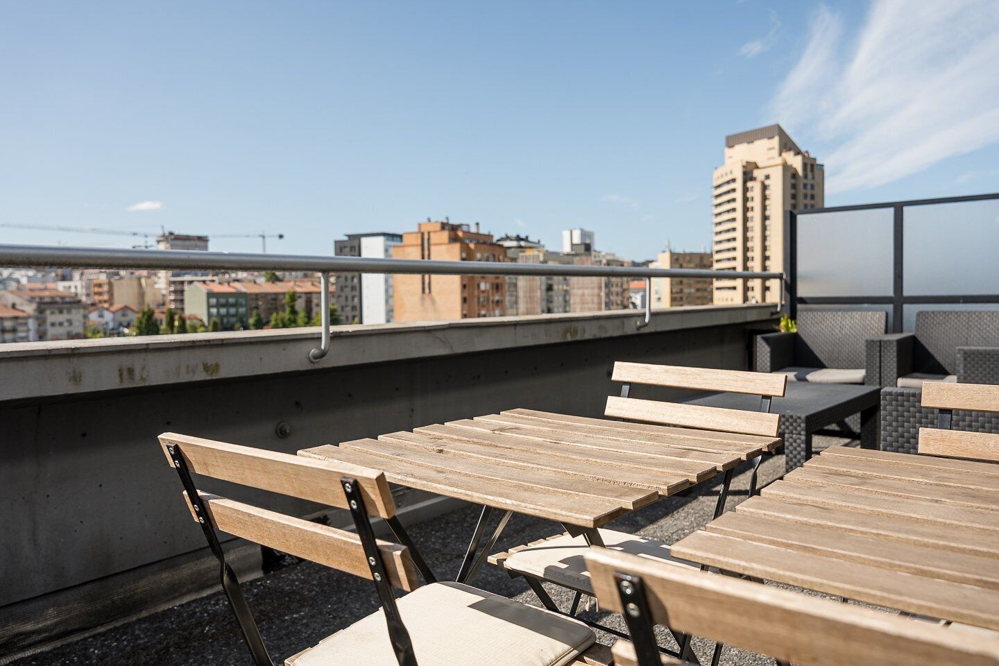 Een terras op het dak met houten tafels en stoelen, een metalen hekwerk en uitzicht over de stad.