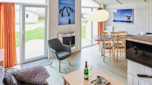 A modern living room with a sofa, a coffee table with a bottle and glass, a fireplace, and a dining area with chairs and a table. - 4 Star Holiday Home in Friedrichkoog (Friedrichskoog)