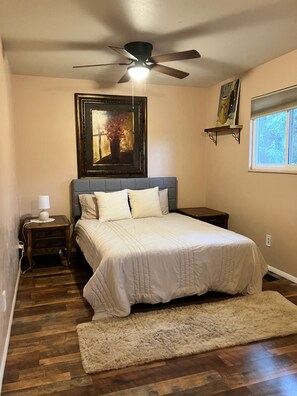 Une chambre à coucher avec un lit, une table de chevet, une lampe, un ventilateur de plafond, un tableau et une fenêtre.