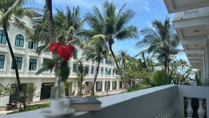 A balcony with a white railing, a vase with a red flower, and a book. In the background, there is a building with multiple windows and palm trees. - NS Home B5-301 (Phu Quoc)