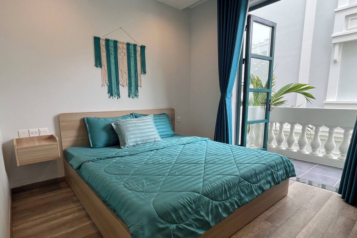 A bedroom with a bed, a bedside table, a wall-mounted macrame decoration, and a view of the outside balcony.