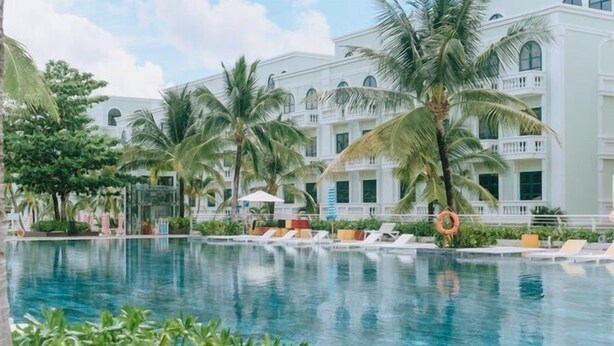 A pool area with lounge chairs and palm trees, surrounded by a large building with multiple balconies. - NS Home B5-301 (Phu Quoc)