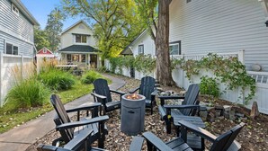 A patio with Adirondack chairs and a fire pit, surrounded by a white fence and trees.