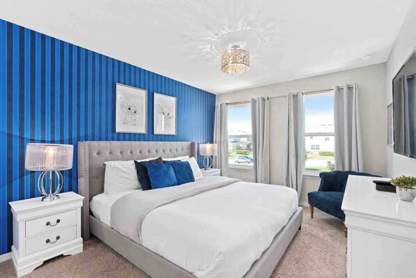 A bedroom with a large bed, a nightstand, a lamp, a chair, and a window with curtains.
