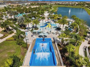 An aerial view of a water park with a large blue water slide, palm trees, and a lake in the background. - Luxury 5BR Disney Retreat w/ Private Pool & Themed Rooms – Steps to Clubhouse! (Kissimmee)