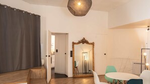 Ein modernes Wohnzimmer mit Holzboden, einem Essbereich mit rundem Tisch und Stühlen, einem großen Spiegel und einer Deckenleuchte.