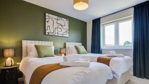 A hotel room with two beds, a large window with curtains, a wall art piece, and a lamp on a bedside table.