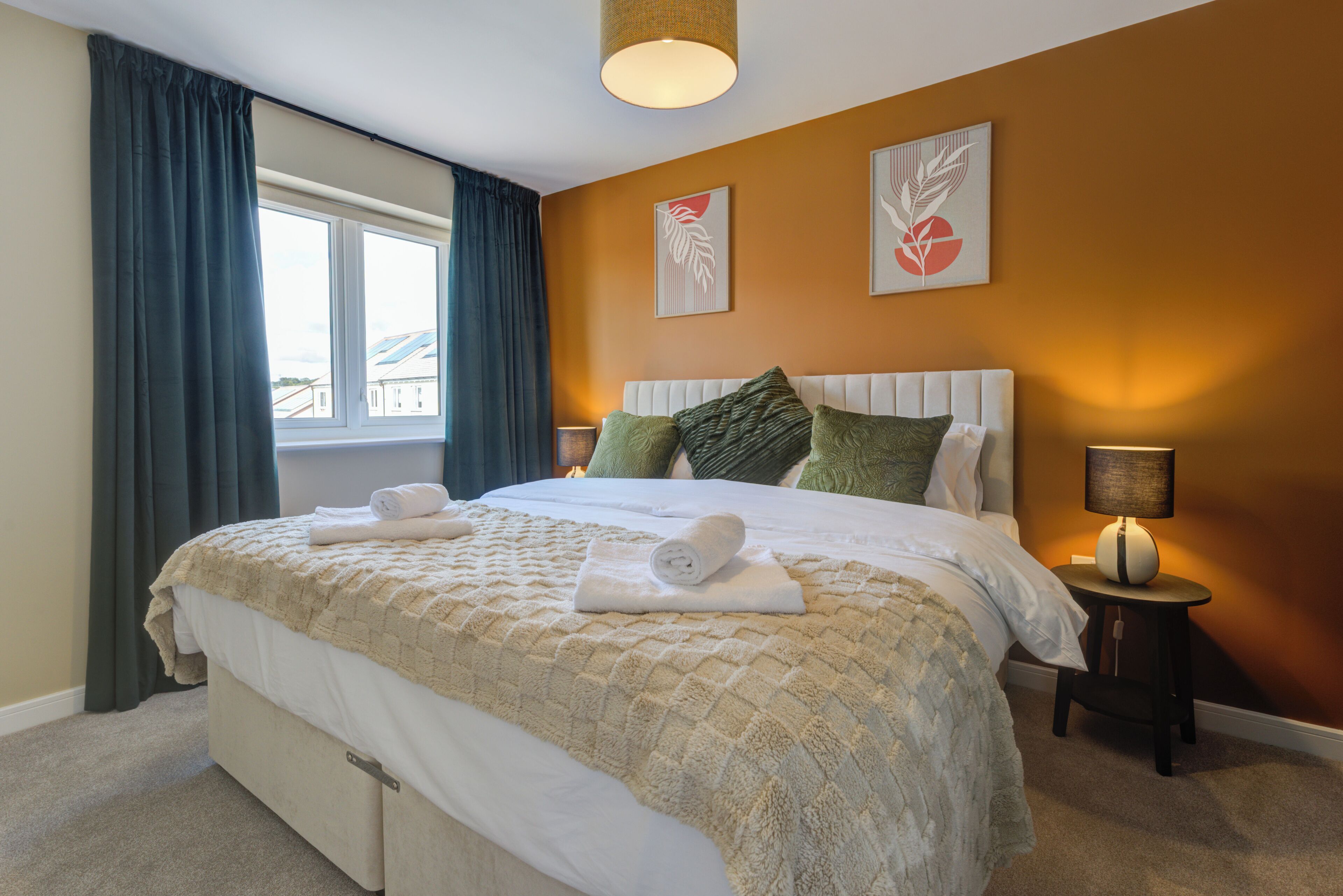 A neatly made bed with a quilt, pillows, and rolled towels. There is a bedside lamp and a small table with a lamp. The room has a window with curtains and artwork on the wall.