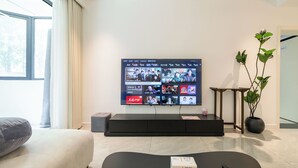 Ein modernes Wohnzimmer mit großem Flachbildfernseher an der Wand, einer Couch, einem Couchtisch, einer Topfpflanze und einem kleinen Tisch mit Fernbedienung.