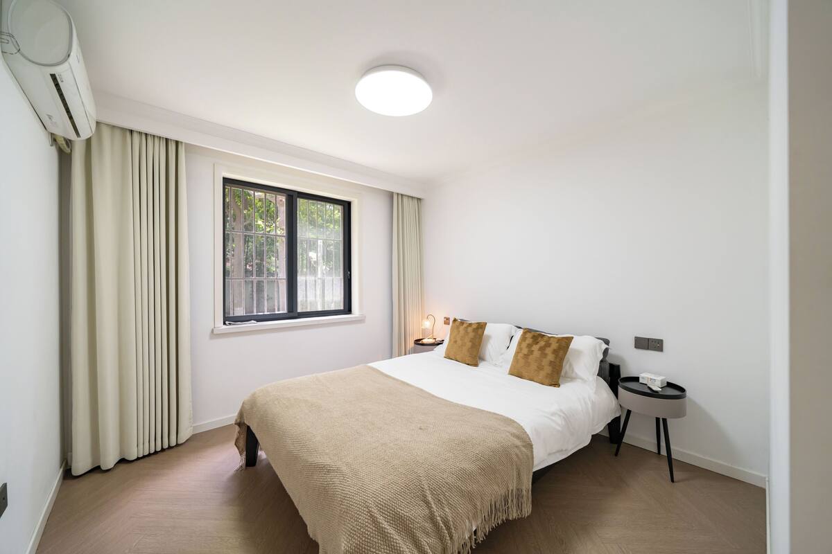 A hotel room with a bed, a bedside table, a window, and an air conditioning unit.