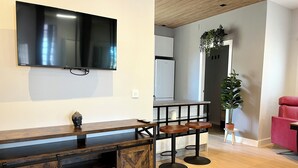 Un salon moderne avec un meuble de rangement en bois pour l’équipement audiovisuel, un téléviseur à écran plat et, au fond, un coin cuisine.
