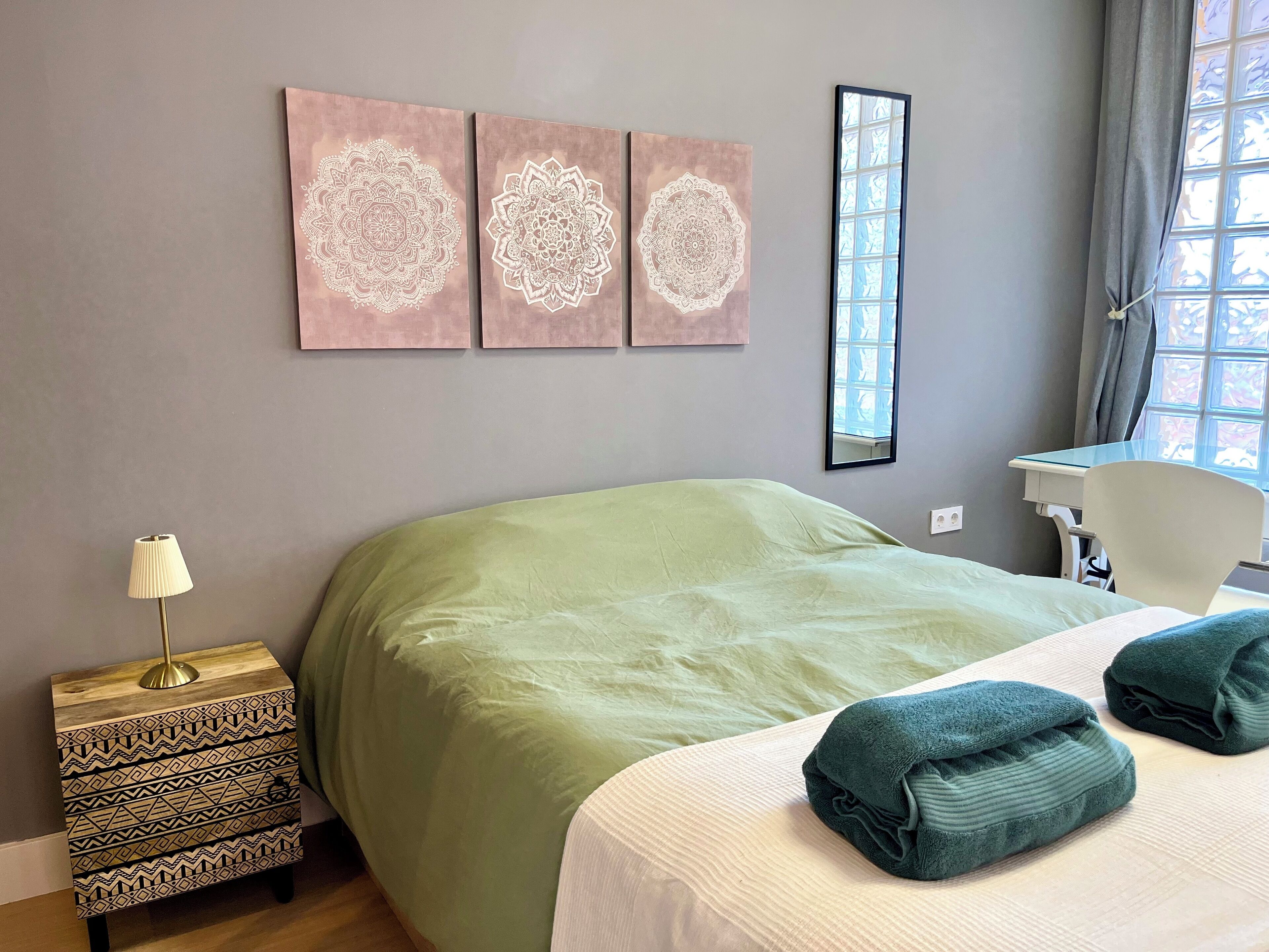 A bedroom with a large bed, a bedside table with a lamp, a desk with a chair, and three wall-mounted artworks.