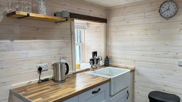 A modern bathroom with wooden panel walls, a wooden countertop, a sink, a mirror, a kettle, and a trash bin. - Cozy Seaside Retreat in Peterhead (Peterhead)