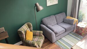 Ein Wohnzimmer mit grüner Wand, einem grauen Sofa, einem karierten Sessel mit gelbem Kissen, einer Stehlampe und einem gerahmten Motivationszitat.