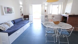 A bright living room with a blue sofa, white dining table, and chairs. There is a fireplace and a window with curtains. - Villetta Blu sul Mare a Fontane Bianche by Wonderful Italy (Syracuse)