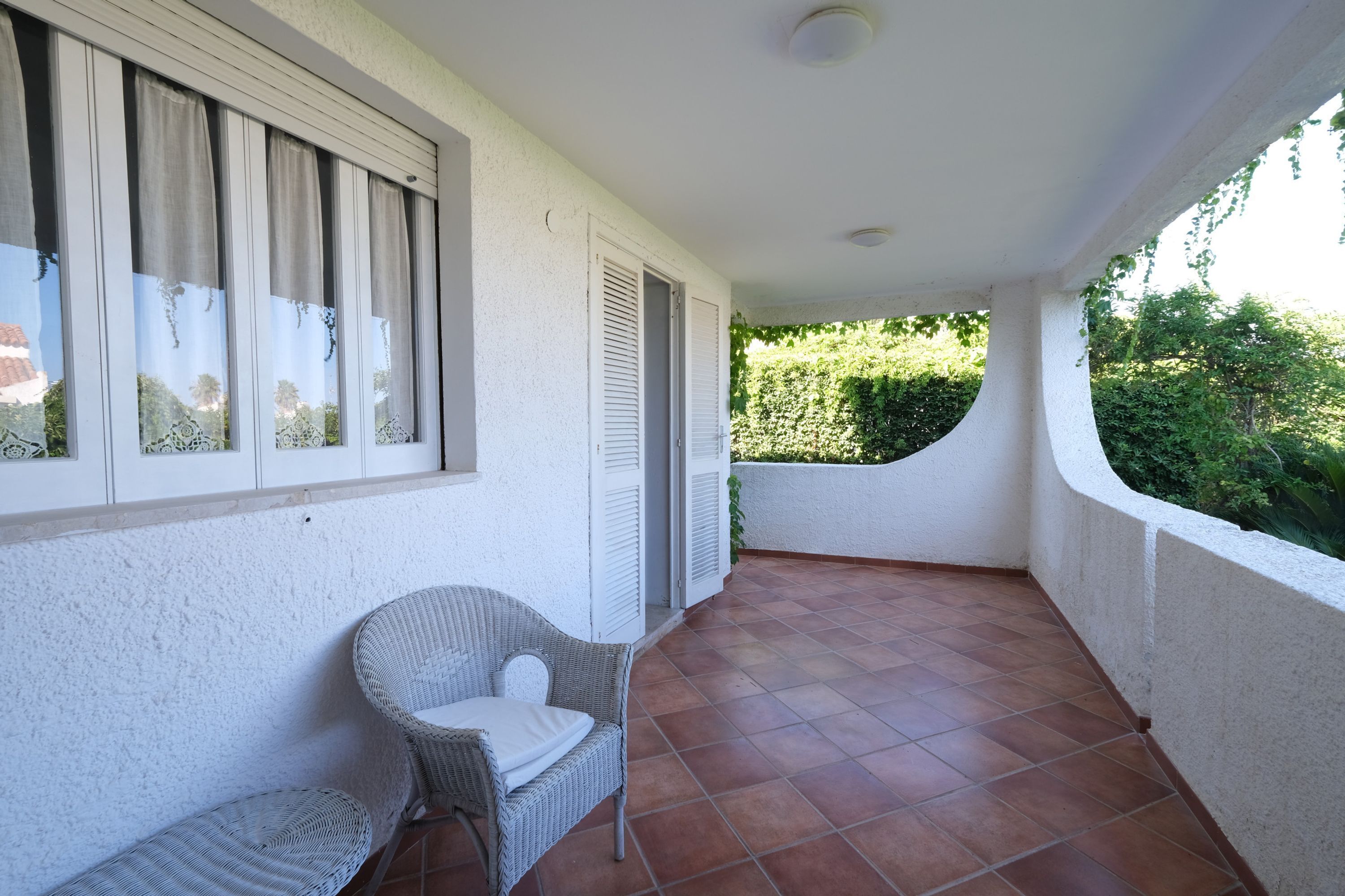 A balcony with a wicker chair, a window with shutters, and a view of greenery.