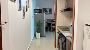 Un couloir étroit mène à une cuisine équipée d’un lave-linge, d’un réfrigérateur et d’un four à micro-ondes. Des tableaux encadrés ornent les murs et une plante en pot se trouve dans le couloir.