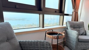 Une pièce moderne avec des fauteuils à revêtement gris, une table d’appoint en osier et une vue sur une étendue d’eau, visible à travers de grandes fenêtres.