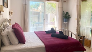 A neatly made bed with a white and red blanket, a metal headboard, and a view of a balcony with plants and a table.