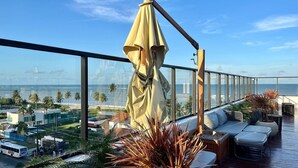 A balcony with a view of the ocean, a large umbrella, and outdoor seating. - Get By Side (João Pessoa)